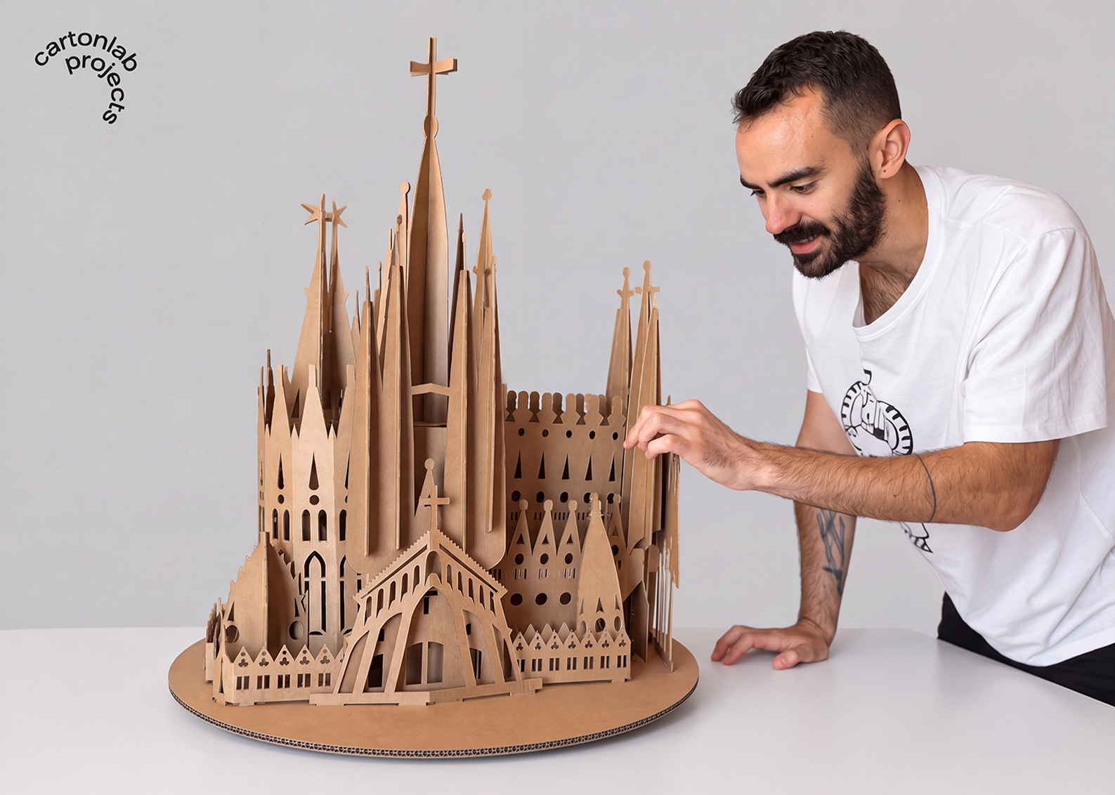 Persona ajustando una pieza de la maqueta de cartón inspirada en la Sagrada Família, sobre una base circular y una mesa blanca